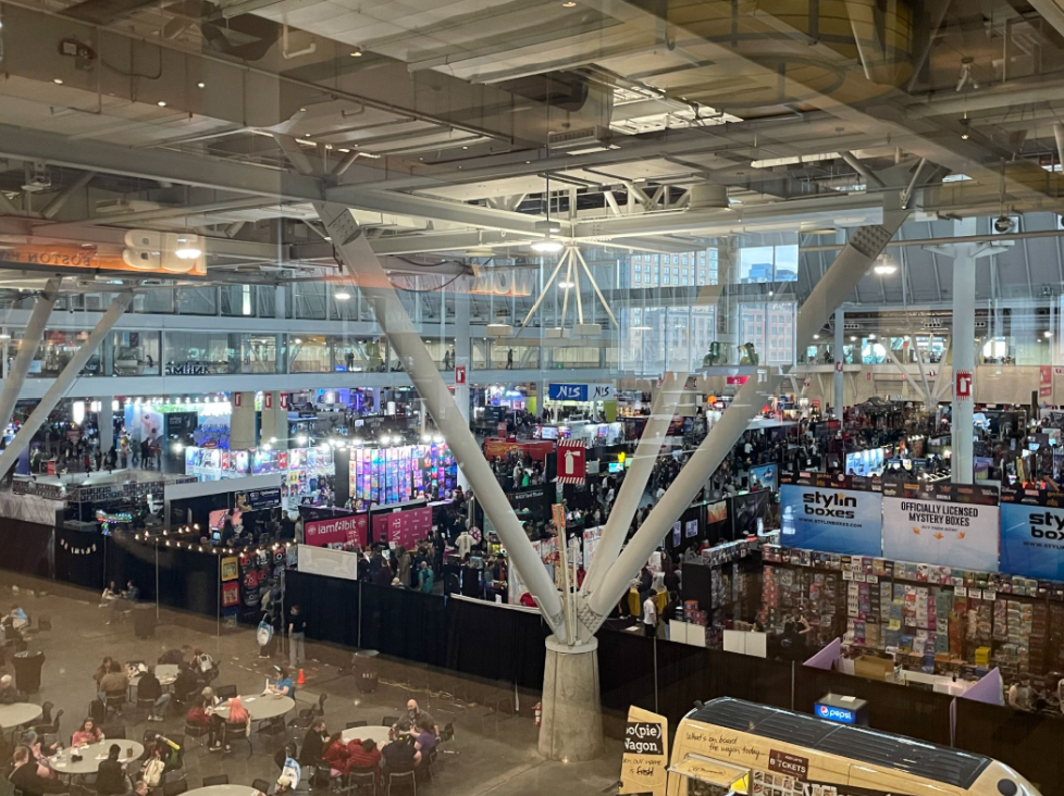 Photo taken from an escalator overlooking the PAX East exposition hall, a large crowded space with tables for table top gaming and booths selling video game merch.