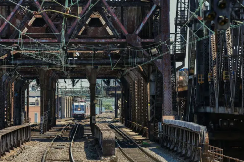 This photo is a web of rusting steal beams and green copper wiring strung over train tracks. A train can be seen approaching in the distance. Everything looks run down.