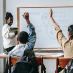 Two students raise their hands in a classroom. Photo courtesy Katerina Holmes on Pexels.