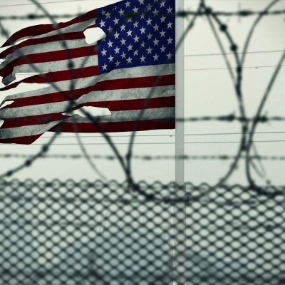 American Flag with holes in it behind a barbed wire fence. Photo courtesy of TayebMEZAHDIA on Pixabay.