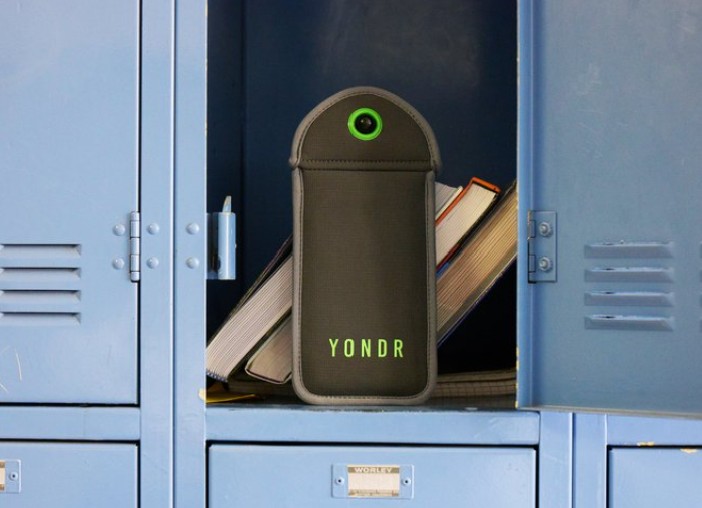 A photo of the Yondr phone pouch, a grey plush case with bright green accents, situated in a blue school locker propped up by books