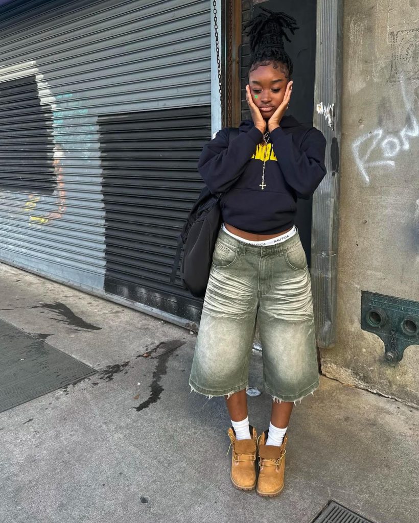 Instagram fashion post of a Black teen girl posing in a pair of acid washed jorts, a black hoodie, and Timberland boots.