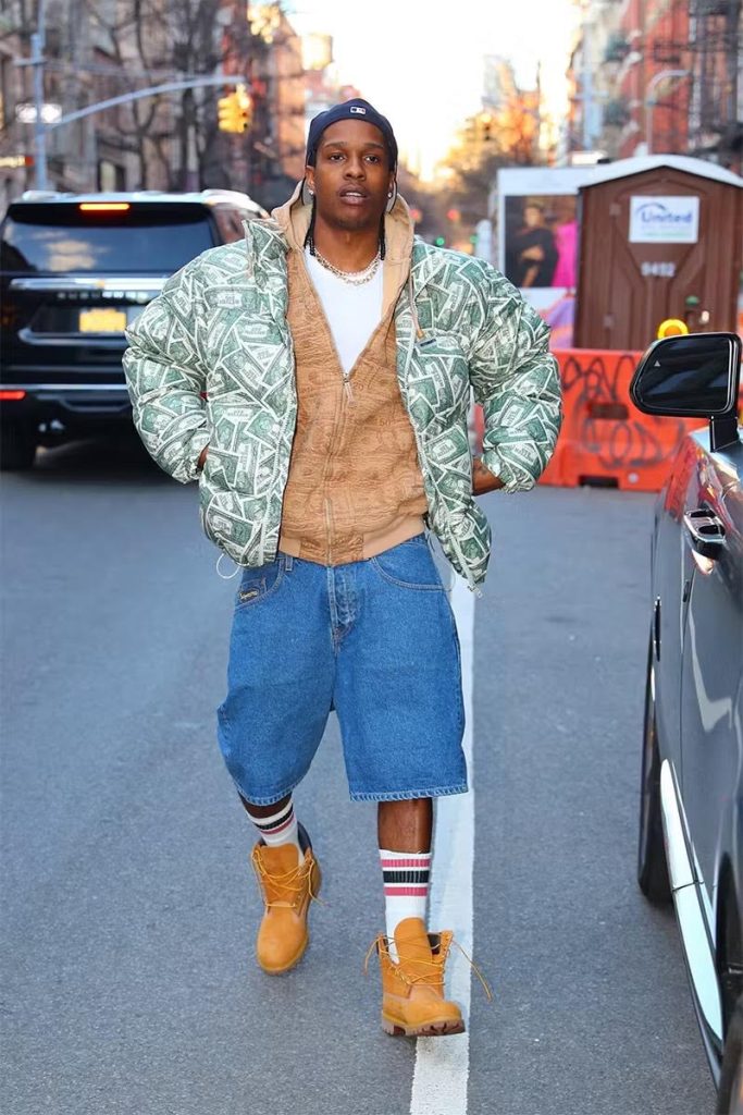 Instagram fashion post of a Black man walking in a city wearing a puffer jacket with dollar bill images printed on it, a brown sweater, blue jorts, and Timberlands.