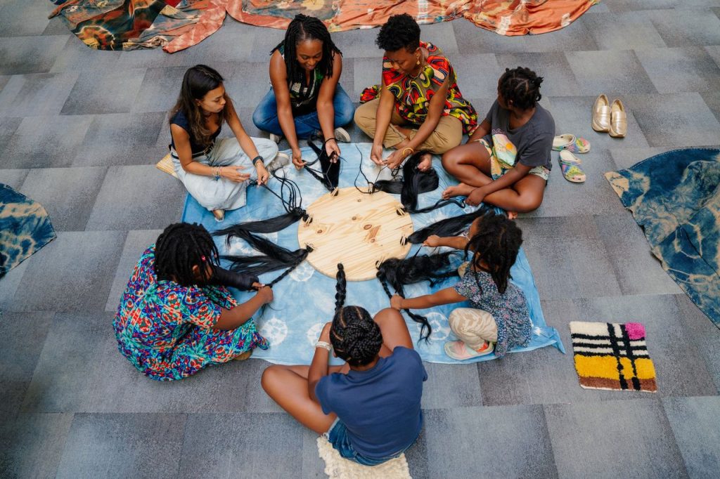 A group of teens and educators sitting in a circle and braiding hair for a multimedia art project.