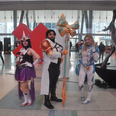 3 cosplayers posing in a convention hall lobby. From left to right is a woman dressed as Syndrome's Star Guardian Skin from League of Legends, a man dressed as Jayce from Arcane, and a woman dressed as Sue Storm from Fantastic Four.