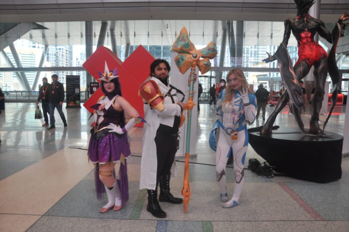 3 cosplayers posing in a convention hall lobby. From left to right is a woman dressed as Syndrome's Star Guardian Skin from League of Legends, a man dressed as Jayce from Arcane, and a woman dressed as Sue Storm from Fantastic Four.