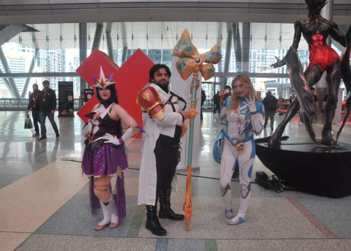 3 cosplayers posing in a convention hall lobby. From left to right is a woman dressed as Syndrome's Star Guardian Skin from League of Legends, a man dressed as Jayce from Arcane, and a woman dressed as Sue Storm from Fantastic Four.