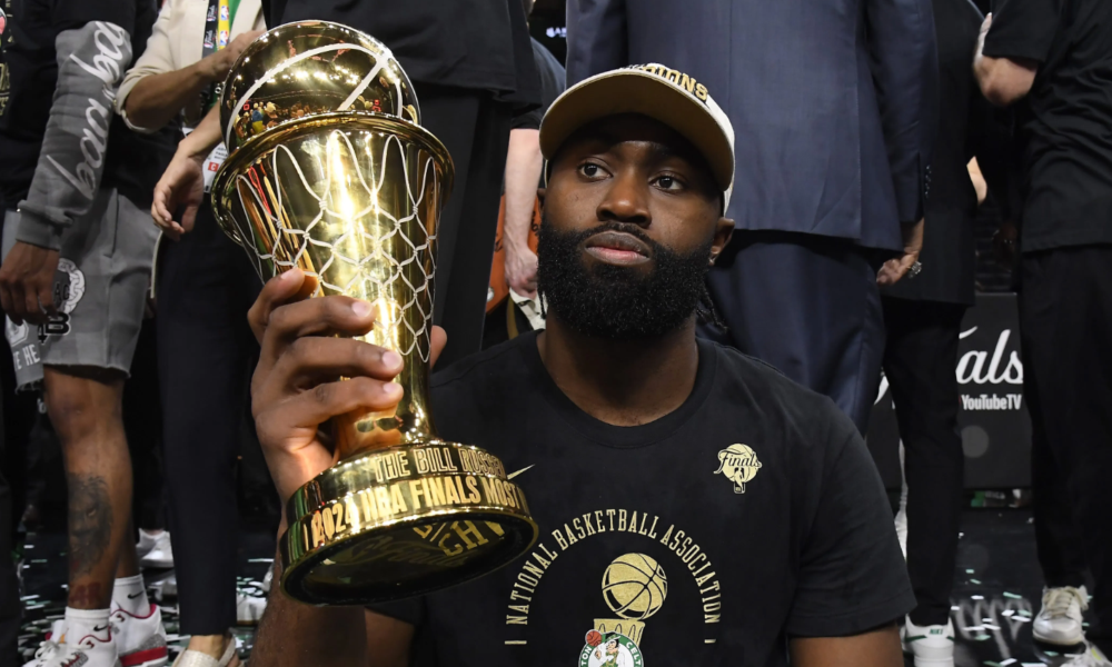 Jaylen Brown sitting courtside and holding a large golden MVP trophy.