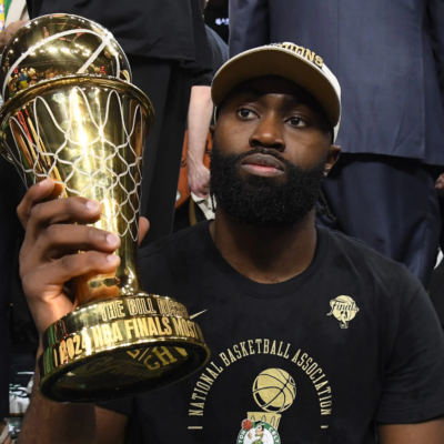 Jaylen Brown sitting courtside and holding a large golden MVP trophy.