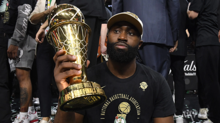Jaylen Brown sitting courtside and holding a large golden MVP trophy.