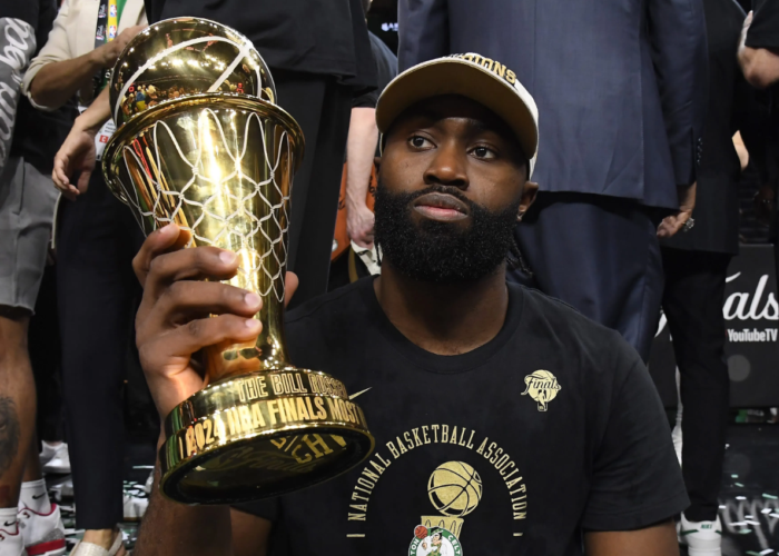 Jaylen Brown sitting courtside and holding a large golden MVP trophy.