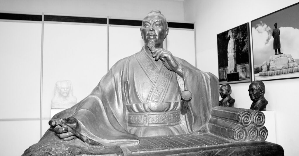 Black and white photo of a statue of Sun Tzu, depicted writing scrolls of strategy.