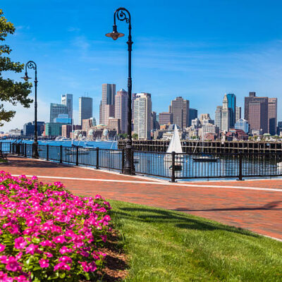 A brick walkway in East Boston's Pier's Park invites you to stroll past the lush green grass and bright flowers to the black wrought-iron railing by the waterfront. Gaze out across the Boston Harbor to see sailboats gliding by or the numerous buildings of all sizes in downtown Boston. With the clear blue sky, lights are not needed, but decorative street lamps are ready for nightfall.