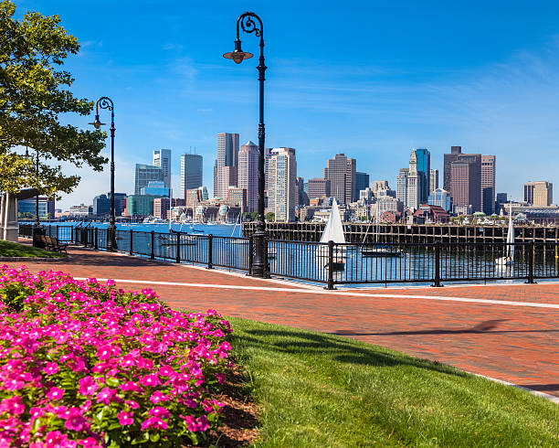 A brick walkway in East Boston's Pier's Park invites you to stroll past the lush green grass and bright flowers to the black wrought-iron railing by the waterfront. Gaze out across the Boston Harbor to see sailboats gliding by or the numerous buildings of all sizes in downtown Boston. With the clear blue sky, lights are not needed, but decorative street lamps are ready for nightfall.